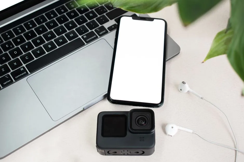 Flat lay of a laptop, smartphone with blank screen, action camera, and earphones on white background.